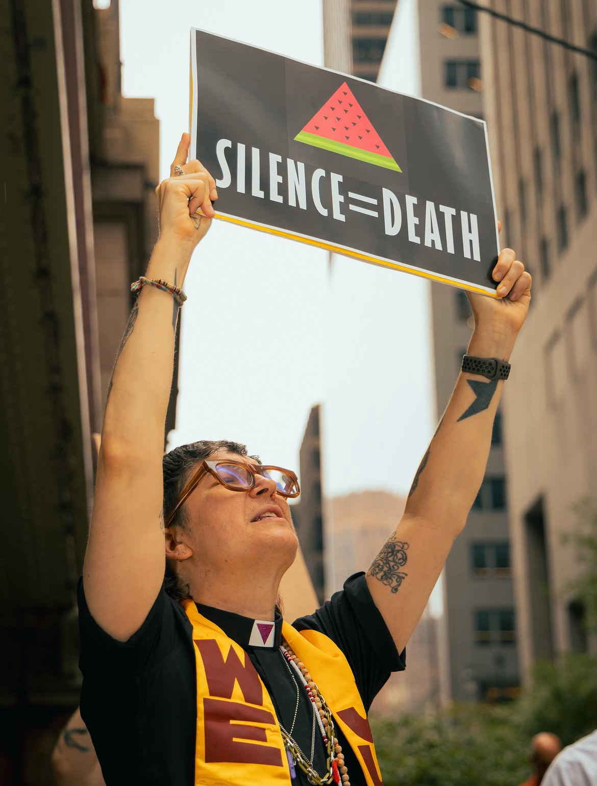 Rev. Elæ Moss Benedetto holding a Silence=Death sign at a protest, wearing a clerical collar and stole
